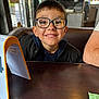 child, boy, glasses, smile, table, indoor, cafe, wooden_table, jacket, person, face, happy, reflection, casual, seated, short_hair, light, background, window, arm