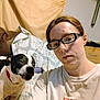 person, dog, glasses, indoor, quilt, couch, cardboard_box, white_shirt, red_collar, pet, brown_hair, face, expression, seated, looking_at_camera, home, casual, furniture, blanket, wall