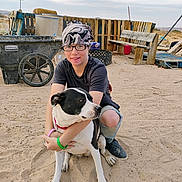 Muffin joined the competition — help win amazing prizes! person, dog, sand, outdoor, glasses, headscarf, bench, wheelbarrow, fence, collar, knee_pad, smile, sitting, pet, desert, sky, clouds, wooden_pallets, shoes, casual_clothing