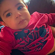 Nico is registered to the contest to win money with this photo: baby, child, face, red_sweater, black_graphic, curious_eyes, cushion, indoor, portrait, young_child, closeup, soft_light, comfortable, casual_clothing, infant, person, cute, looking_at_camera, seated, relaxed