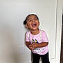 child, toddler, laughing, smile, teeth, standing, pink_shirt, backpack_straps, black_shorts, braided_hair, hair_buns, indoor, white_wall, hardwood_floor, doorway, candid, joyful, portrait, sneakers, playful