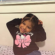 Janae is registered to the contest to win money with this photo: bed, braided_hair, casual_clothing, child, cute, girl, happy, hello_kitty, indoor, patterned_sheet, person, pink_bow, playful, portrait, seated, sheet, smiling, sweater, wall, young