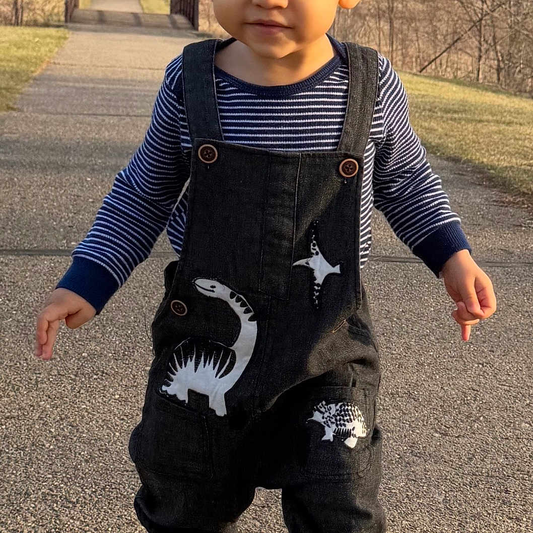 Dalthon is registered to the contest to win money with this photo: toddler, child, walking, outdoor, path, overalls, dinosaur, sneakers, park, trees, bridge, sunlight, smiling, person, nature, casual_clothing, daytime, cute, young_child, fashion