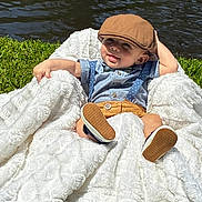 Nikolas is registered to the contest to win money with this photo: baby, child, blanket, outdoor, grass, water, hat, cap, shirt, suspenders, shorts, footwear, cute, relaxed, sunlight, nature, portrait, smile, baby_boy, fashion