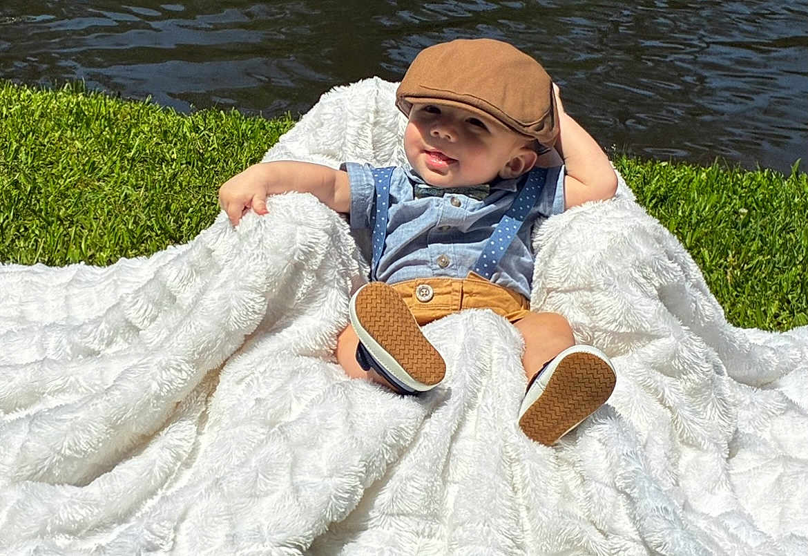 Nikolas is registered to the contest to win money with this photo: baby, child, blanket, outdoor, grass, water, hat, cap, shirt, suspenders, shorts, footwear, cute, relaxed, sunlight, nature, portrait, smile, baby_boy, fashion