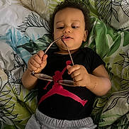 Lyam participe au concours pour gagner de l'argent avec cette photo : bed, bedding, black_shirt, blanket, casual, child, clothing, curious, curly_hair, eyeglasses, face, gray_pants, hand, indoor, laying_down, leaf_pattern, person, red_logo, toddler, young_child