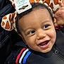 toddler, child, smiling, face, headband, giraffe_pattern, clothing, black_jacket, red_stripe, blue_stripe, white_stripe, hand, happy, cute, indoors, person, closeup, expression, young_child, playful