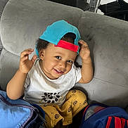 Lyam participe au concours pour gagner de l'argent avec cette photo : toddler, child, smiling, cap, backward_cap, sitting, couch, white_tshirt, yellow_pants, patterned_pants, indoor, playful, happy, person, young_child, bag, colorful, cute, portrait, casual