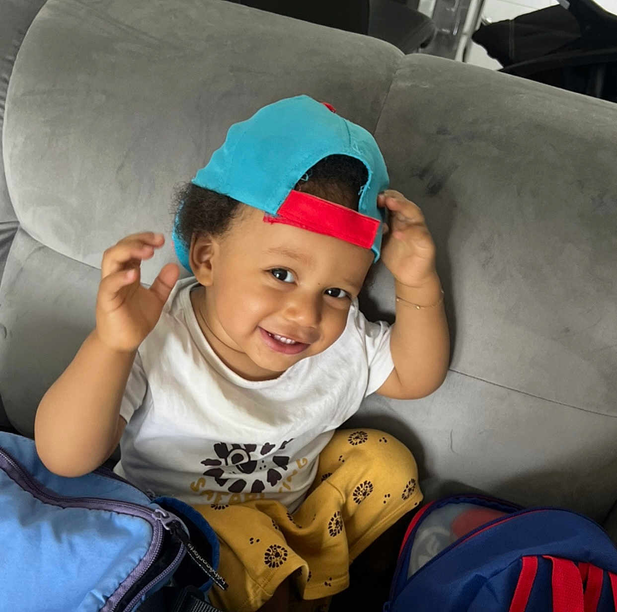 Lyam participe au concours pour gagner de l'argent avec cette photo : toddler, child, smiling, cap, backward_cap, sitting, couch, white_tshirt, yellow_pants, patterned_pants, indoor, playful, happy, person, young_child, bag, colorful, cute, portrait, casual
