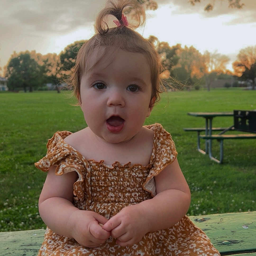 Jadynn joined the competition — help win amazing prizes! baby, child, cute, dress, expression, floral_pattern, grass, green, hair_clip, nature, orange, outdoor, park, person, picnic_table, pony_tail, sitting, sunset, toddler, trees