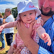 Amelia is registered to the contest to win money with this photo: baby, child, man, hat, outdoor, sunny, people, family, holding, smiling, blue_shirt, beard, grass, cloudy_sky, patterned_clothing, casual, daytime, portrait, happy, event