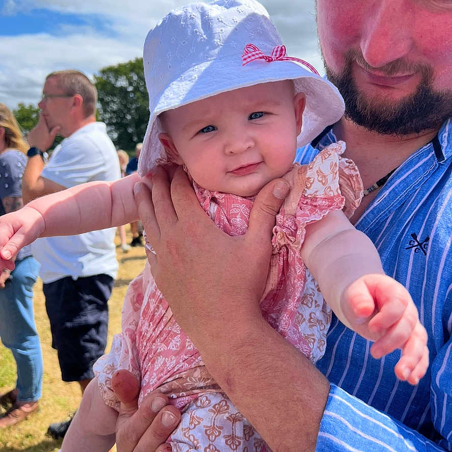 Amelia is registered to the contest to win money with this photo: baby, beard, blue_shirt, casual, child, cloudy_sky, daytime, event, family, grass, happy, hat, holding, man, outdoor, patterned_clothing, people, portrait, smiling, sunny