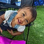 child, toddler, smiling, happy, face, hair, clothing, denim_overalls, chair, outdoor, grass, bag, pink, people, background, daylight, playful, portrait, person, fun