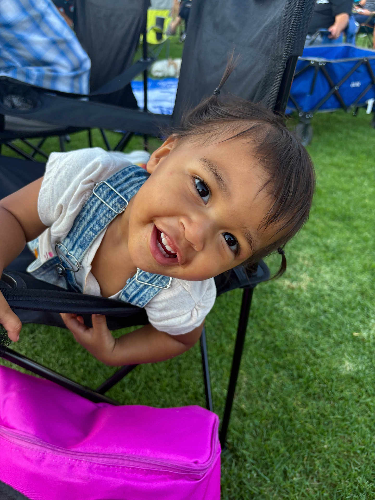 Zia is registered to the contest to win money with this photo: child, toddler, smiling, happy, face, hair, clothing, denim_overalls, chair, outdoor, grass, bag, pink, people, background, daylight, playful, portrait, person, fun