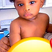 Ethan is registered to the contest to win money with this photo: child, toddler, baby, portrait, afro_hair, curly_hair, big_eyes, bare_torso, kitchen, stove, microwave, cabinets, close_up, neutral_expression, adult_hand, emoji_overlay, indoor, skin, photography, holding