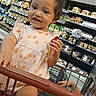 Zoey is registered to the contest to win money with this photo: aisle, casual, child, curious, cute, face, food_packages, grocery_store, hand, holding, indoor, infant, person, polka_dot_dress, product_display, retail, shopping_cart, store_shelf, toddler, young_child