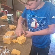 Colten is registered to the contest to win money with this photo: child, boy, cheese, food, table, bread, beverage, cans, kitchen, indoor, casual, blue_shirt, cutting, hands, standing, floor, furniture, snack, meal, contemporary