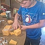 child, boy, cheese, food, table, bread, beverage, cans, kitchen, indoor, casual, blue_shirt, cutting, hands, standing, floor, furniture, snack, meal, contemporary