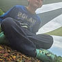child, boy, sitting, cross_legged, blue_shirt, green_shoes, playground, wood_chips, outdoor, shade_cloth, cloudy_sky, casual_clothing, young, daylight, nature, relaxing, leisure, expression, park, footwear