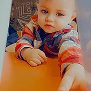 Danaël participe au concours pour gagner de l'argent avec cette photo : toddler, child, table, reaching, curious, indoor, plaid_shirt, face, hand, expression, person, background_art, light, skin, hair, sitting, closeup, focus, colorful, young