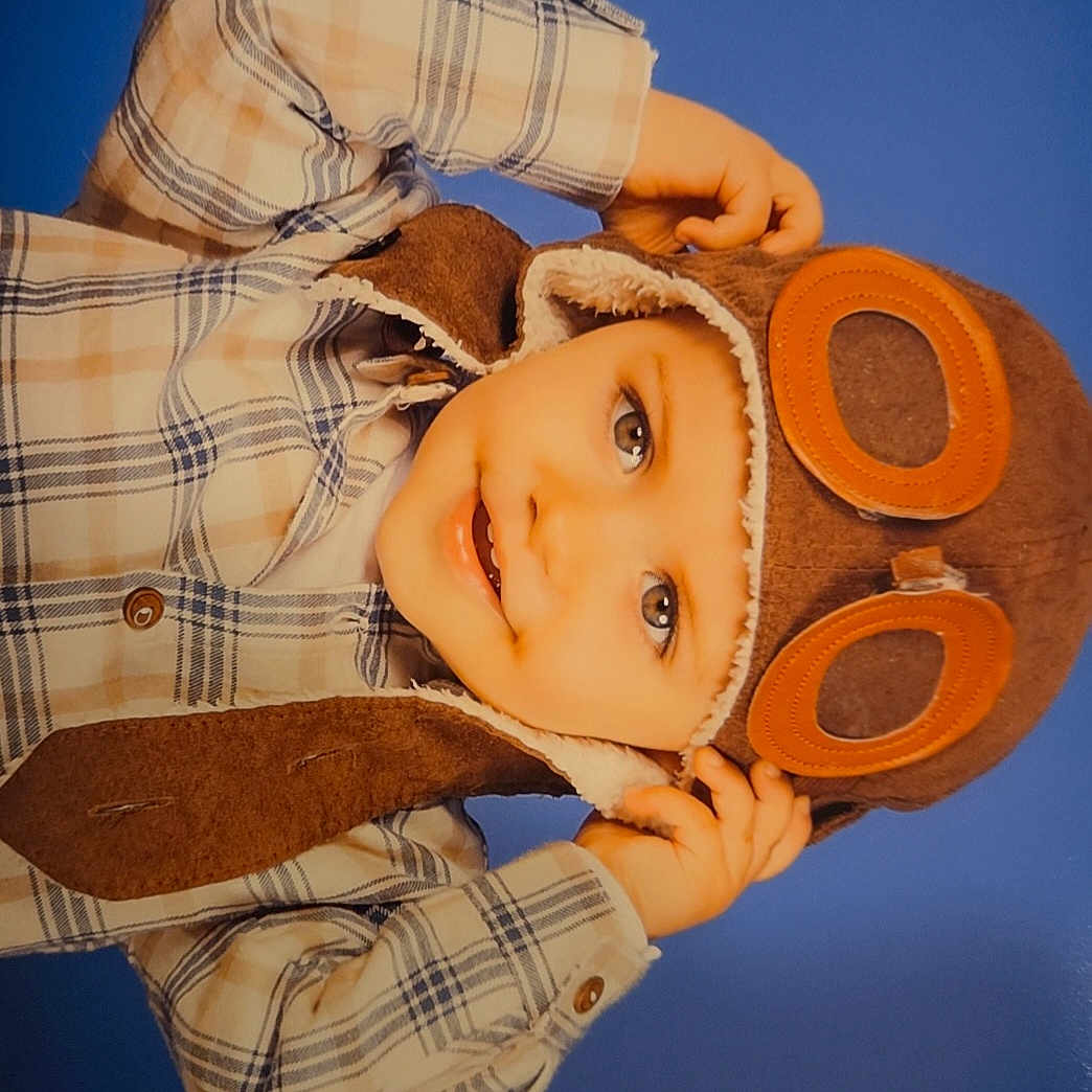 Danaël a rejoint le concours — aidez-le/la à gagner de superbes lots ! aviator_hat, baby, blue_background, casual, child, cozy_clothing, cute, face, fashion, goggles, hands_up, happy, headwear, indoor, person, plaid_shirt, portrait, smile, toddler, warm