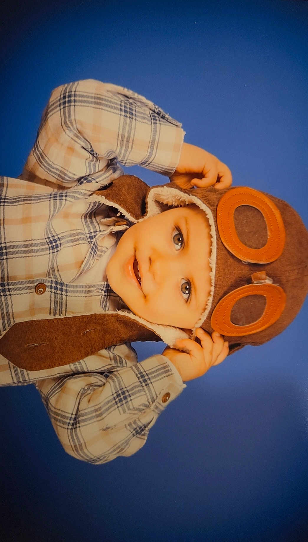 Danaël a rejoint le concours — aidez-le/la à gagner de superbes lots ! child, toddler, aviator_hat, goggles, plaid_shirt, smile, portrait, blue_background, cozy_clothing, hands_up, happy, cute, face, headwear, casual, indoor, baby, fashion, person, warm