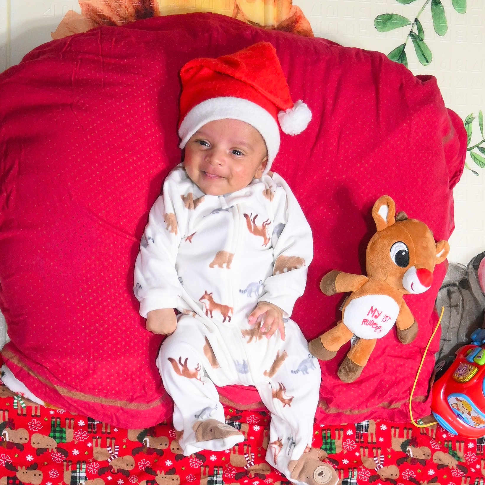 Skanda is registered to the contest to win money with this photo: baby, blanket, celebration, child, christmas, cute, festive, holiday, indoors, infant, lying_down, pajamas, plush_toy, red_cushion, reindeer, santa_hat, sleepwear, smiling, soft_toy, toy_car