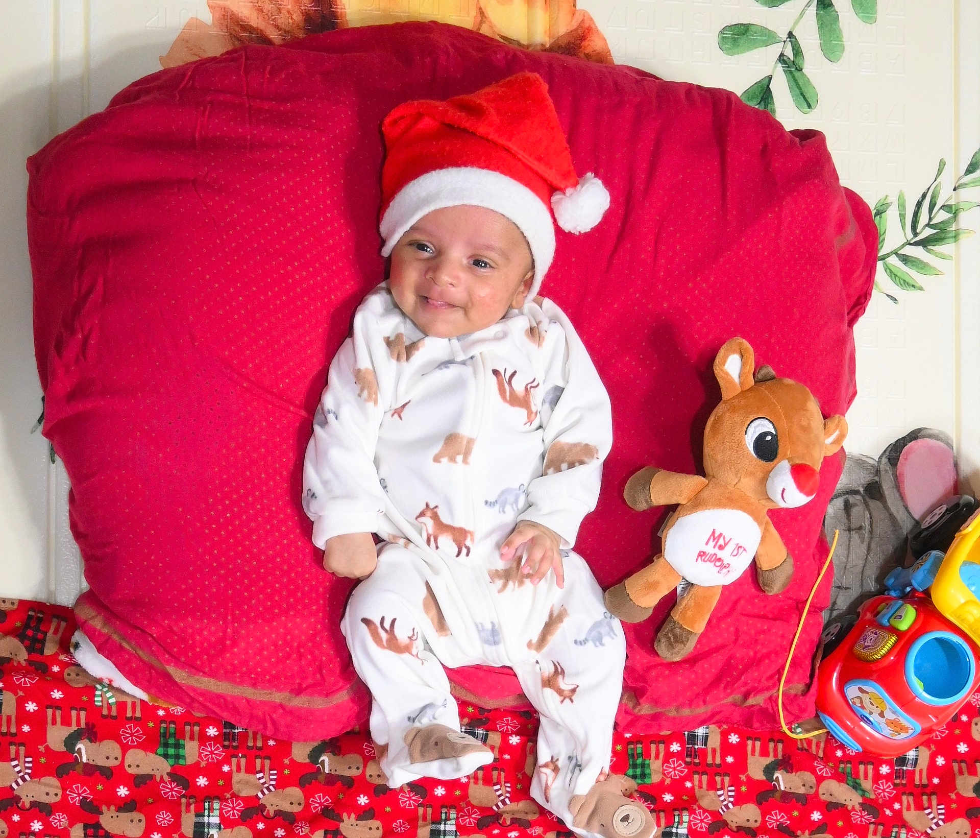 Skanda is registered to the contest to win money with this photo: baby, blanket, celebration, child, christmas, cute, festive, holiday, indoors, infant, lying_down, pajamas, plush_toy, red_cushion, reindeer, santa_hat, sleepwear, smiling, soft_toy, toy_car
