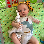Axel a rejoint le concours — aidez-le/la à gagner de superbes lots ! baby, infant, dinosaur_onesie, play_mat, toy, soft_toy, animal_illustrations, curious, lying_down, child, cute, colorful, fabric, clothing, hand, foot, face, indoors, comfort, playtime