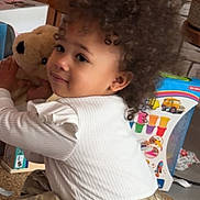 Rym participe au concours pour gagner de l'argent avec cette photo : toddler, child, curly_hair, white_top, golden_skirt, teddy_bear, plush_toy, indoor, basket, toy_box, floor, person, cute, smile, ribbon, hair_accessory, play, cozy, young_child, portrait