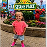 child, girl, toddler, smiling, outdoors, theme_park, sesame_place,  amusement_park, sign, elmo, big_bird, abby_cadabby, flowers, garden, playground, rides, pink_shirt, denim_shorts, pink_shoes, portrait