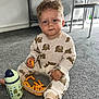 toddler, child, pajamas, bear_pattern, snack, cheese_puffs, plastic_container, sippy_cup, carpet, indoor, messy_hair, barefoot, floor, furniture, table, chair, playpen, drinking_cup, person, cute