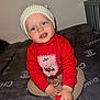 baby, blanket, child, clothing, cozy, cute, face, footwear, happy, hat, holiday, indoor, knit_hat, luxury_brand, portrait, red_sweater, sitting, smiling, toddler, young_child