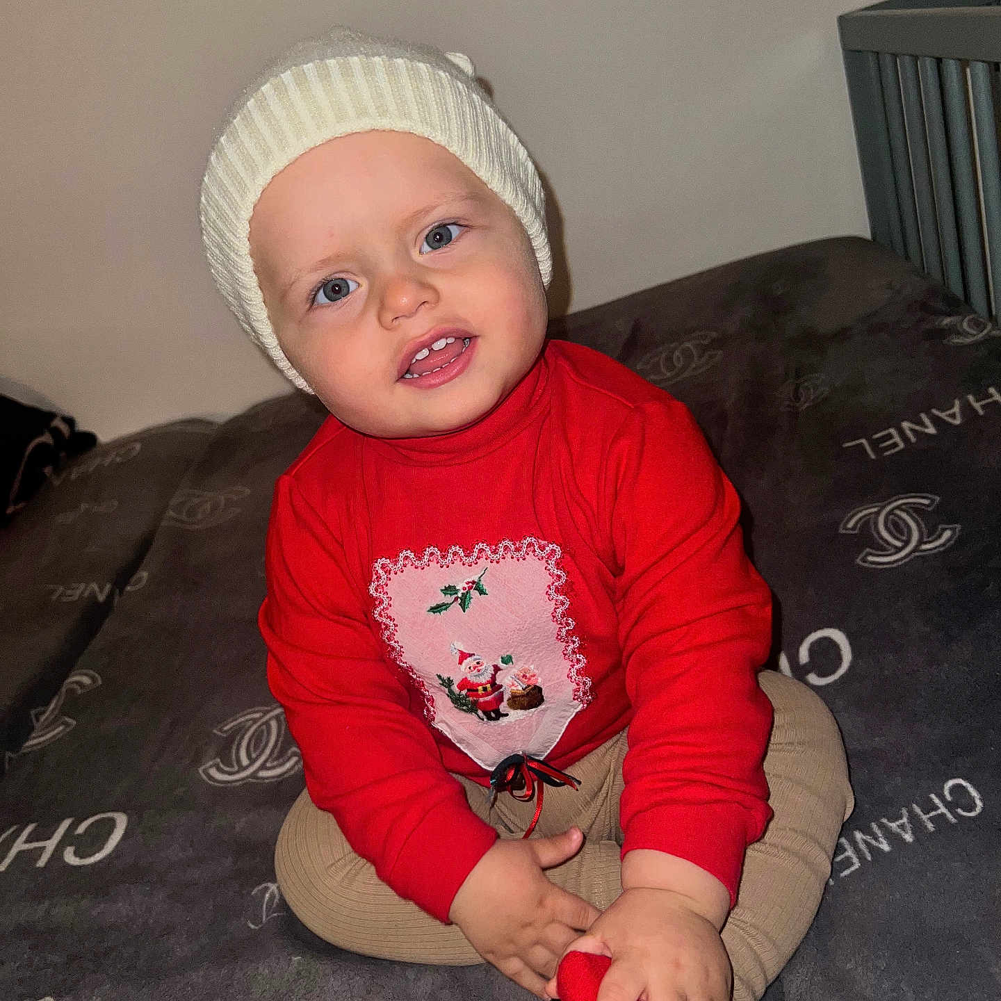 Sami is registered to the contest to win money with this photo: baby, blanket, child, clothing, cozy, cute, face, footwear, happy, hat, holiday, indoor, knit_hat, luxury_brand, portrait, red_sweater, sitting, smiling, toddler, young_child