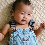 Mateo is registered to the contest to win money with this photo: baby, child, denim_overalls, winking, smiling, face, head, infant, lying_down, textured_blanket, blue_clothing, cute, portrait, skin, arms, one_eye_closed, happy, indoors, person, young_child