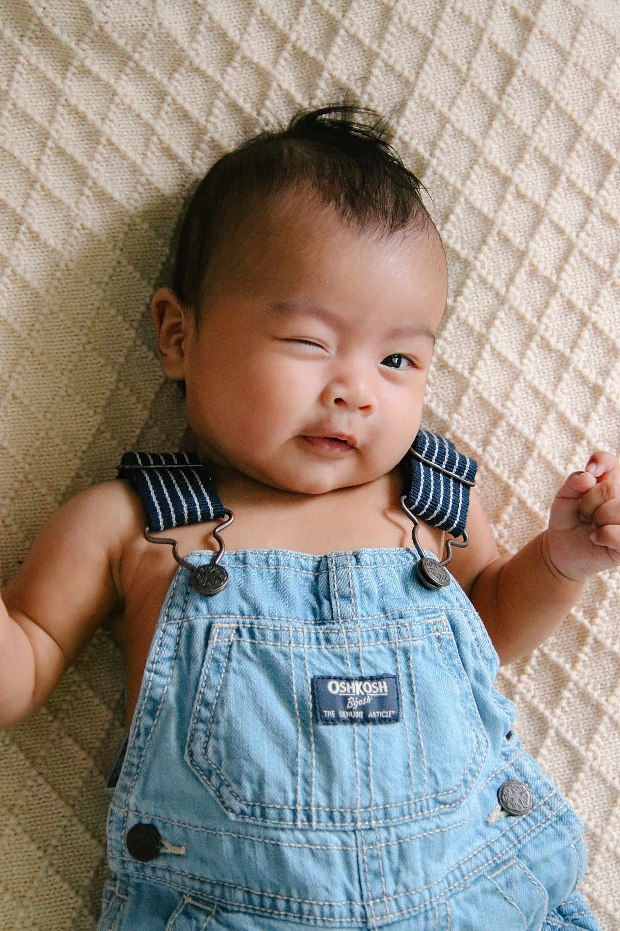Mateo is registered to the contest to win money with this photo: baby, child, denim_overalls, winking, smiling, face, head, infant, lying_down, textured_blanket, blue_clothing, cute, portrait, skin, arms, one_eye_closed, happy, indoors, person, young_child