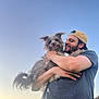man, dog, pet, outdoor, sunset, sky, smiling, cap, beard, casual_clothing, happy, holding, fluffy_dog, tongue_out, friendship, nature, human, animal, portrait, leash