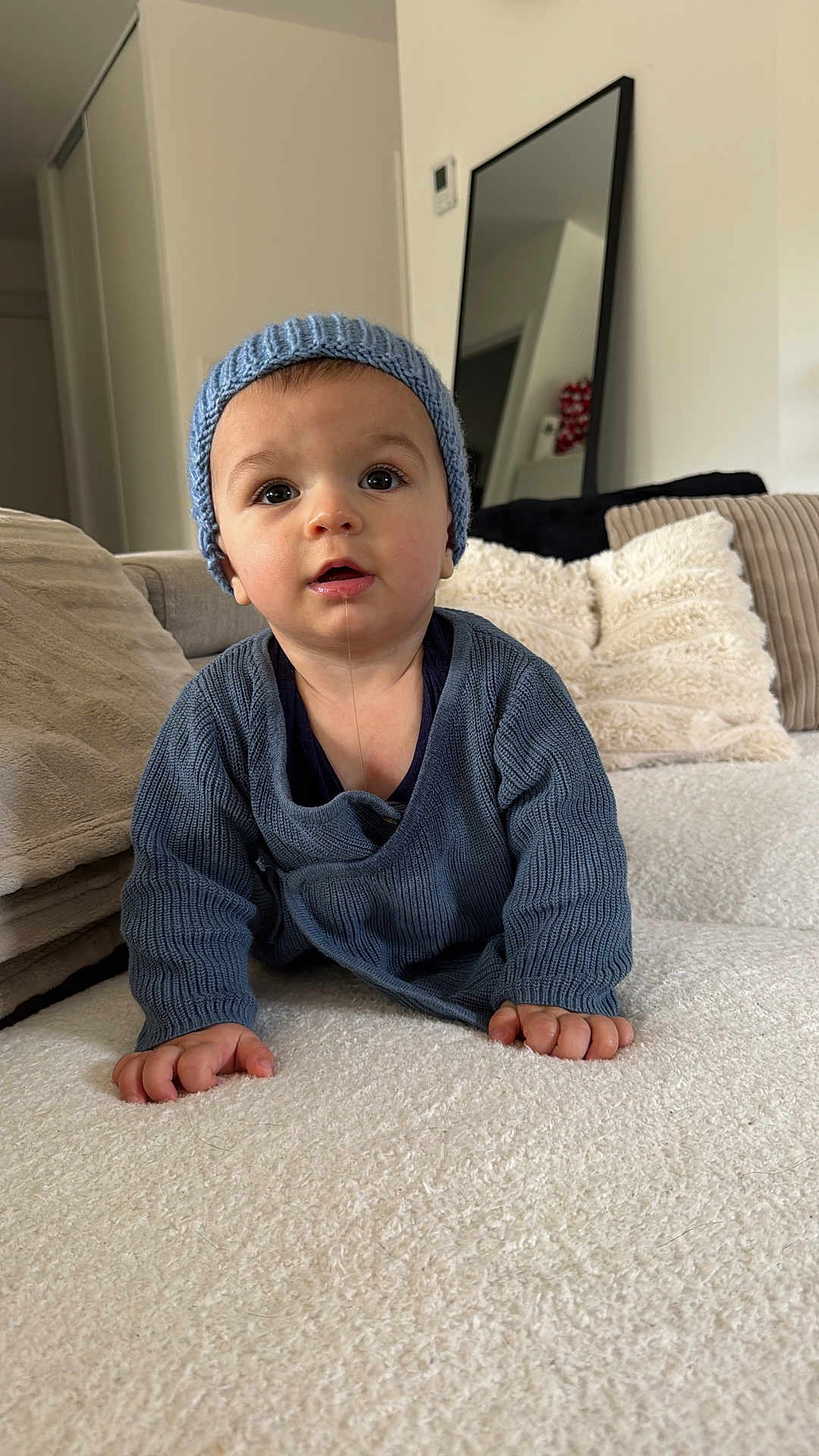 éli a rejoint le concours — aidez-le/la à gagner de superbes lots ! baby, child, crawling, blue_hat, blue_sweater, drool, indoor, cushions, mirror, soft_texture, curious_expression, hands, face, headwear, knitwear, person, portrait, cozy, home, furniture