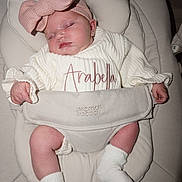 Arabella is registered to the contest to win money with this photo: baby, sleeping, headband, bow, white_sweater, embroidery, name, arabella, cushioned_seat, infant, sock, soft_fabric, hands, legs, pink, comfortable, child, resting, cute, indoors