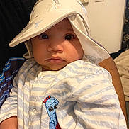 Zaion is registered to the contest to win money with this photo: baby, child, hat, onesie, striped_clothing, embroidered_character, indoor, person, portrait, cute, wide_eyes, hand, adult_arm, fabric, soft_lighting, close_up, face, expression, cozy, infant