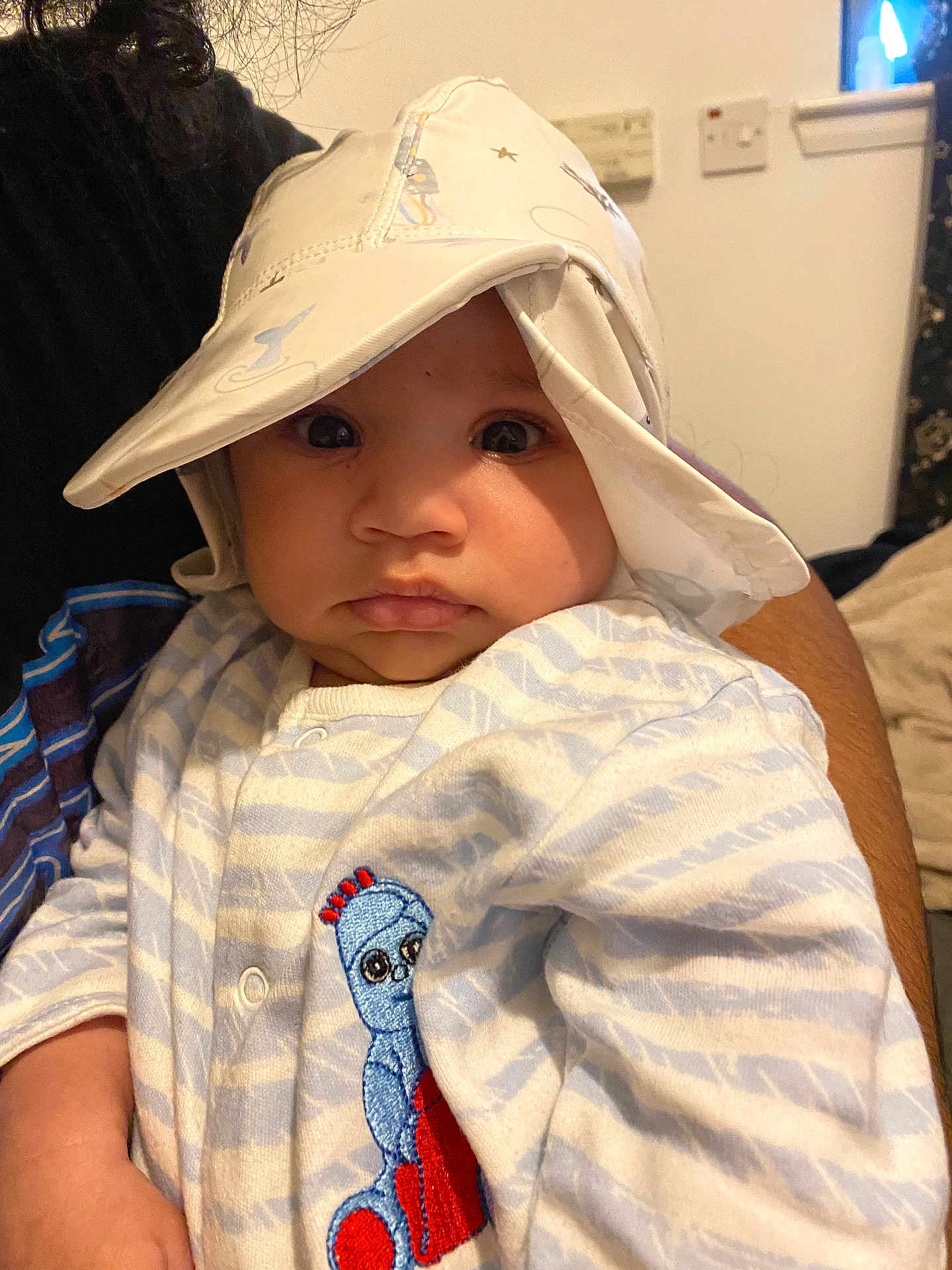 Zaion is registered to the contest to win money with this photo: baby, child, hat, onesie, striped_clothing, embroidered_character, indoor, person, portrait, cute, wide_eyes, hand, adult_arm, fabric, soft_lighting, close_up, face, expression, cozy, infant