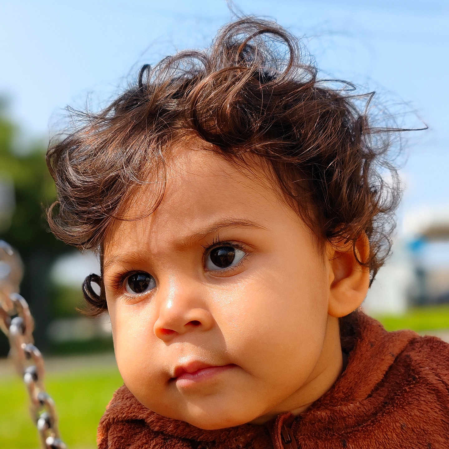 Aïlyne participe au concours pour gagner de l'argent avec cette photo : brown_jacket, casual_clothing, child, childhood, closeup, curious, curly_hair, cute, daylight, expression, face, outdoor, park, person, playground, portrait, sunny, swing, toddler, young_child