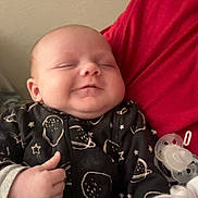 Flynn is registered to the contest to win money with this photo: baby, infant, smile, thumbs_up, blanket, onesie, space_pattern, pacifier, hand, person, red_shirt, indoor, sleepy, cute, face, chubby_cheeks, cozy, child, newborn, portrait
