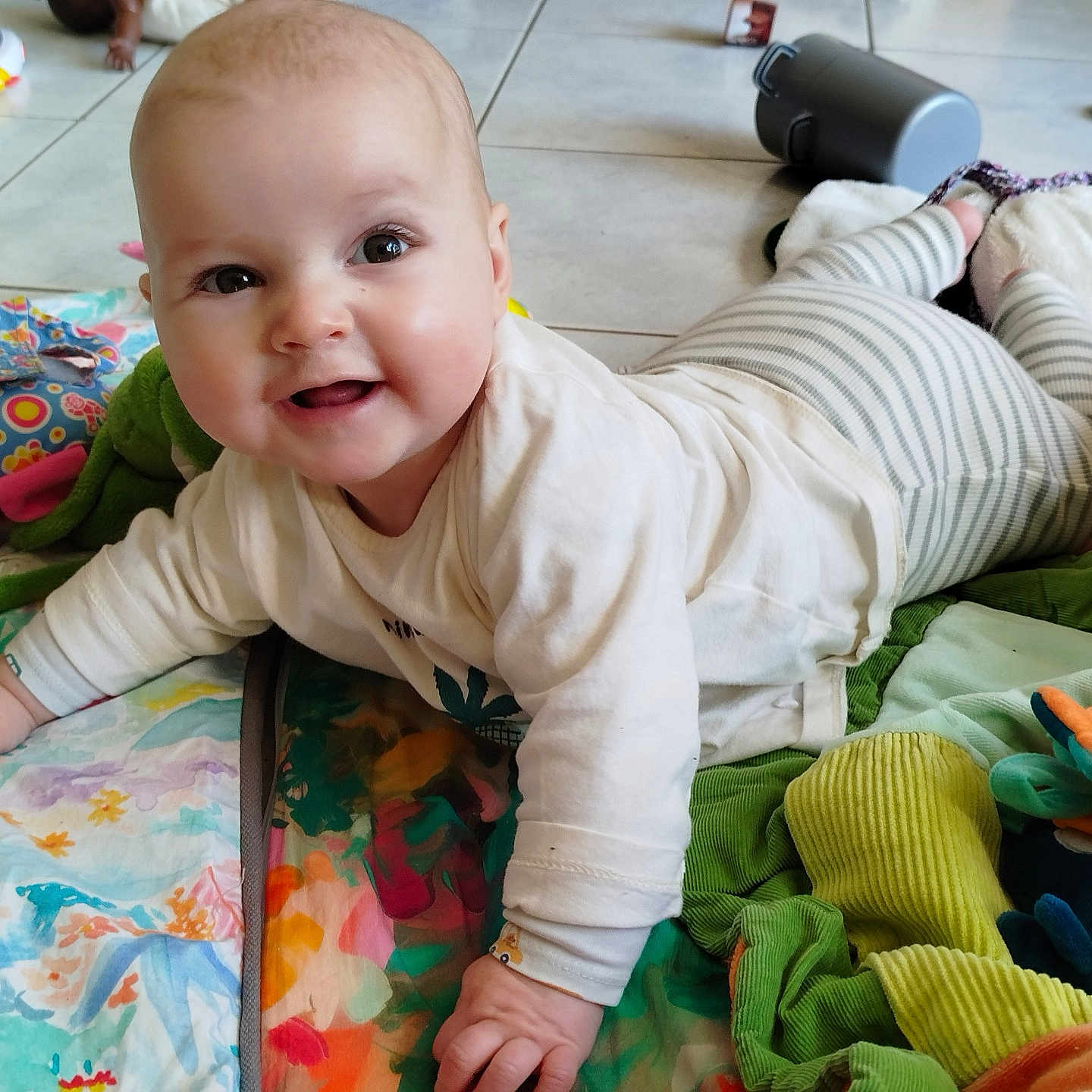 Maddie a rejoint le concours — aidez-le/la à gagner de superbes lots ! baby, smiling, colorful_blanket, toy, floor, tiled_floor, striped_pants, long_sleeve_shirt, indoor, child, happy, cute, playful, infant, person, soft_toys, crawling, bright, face, portrait