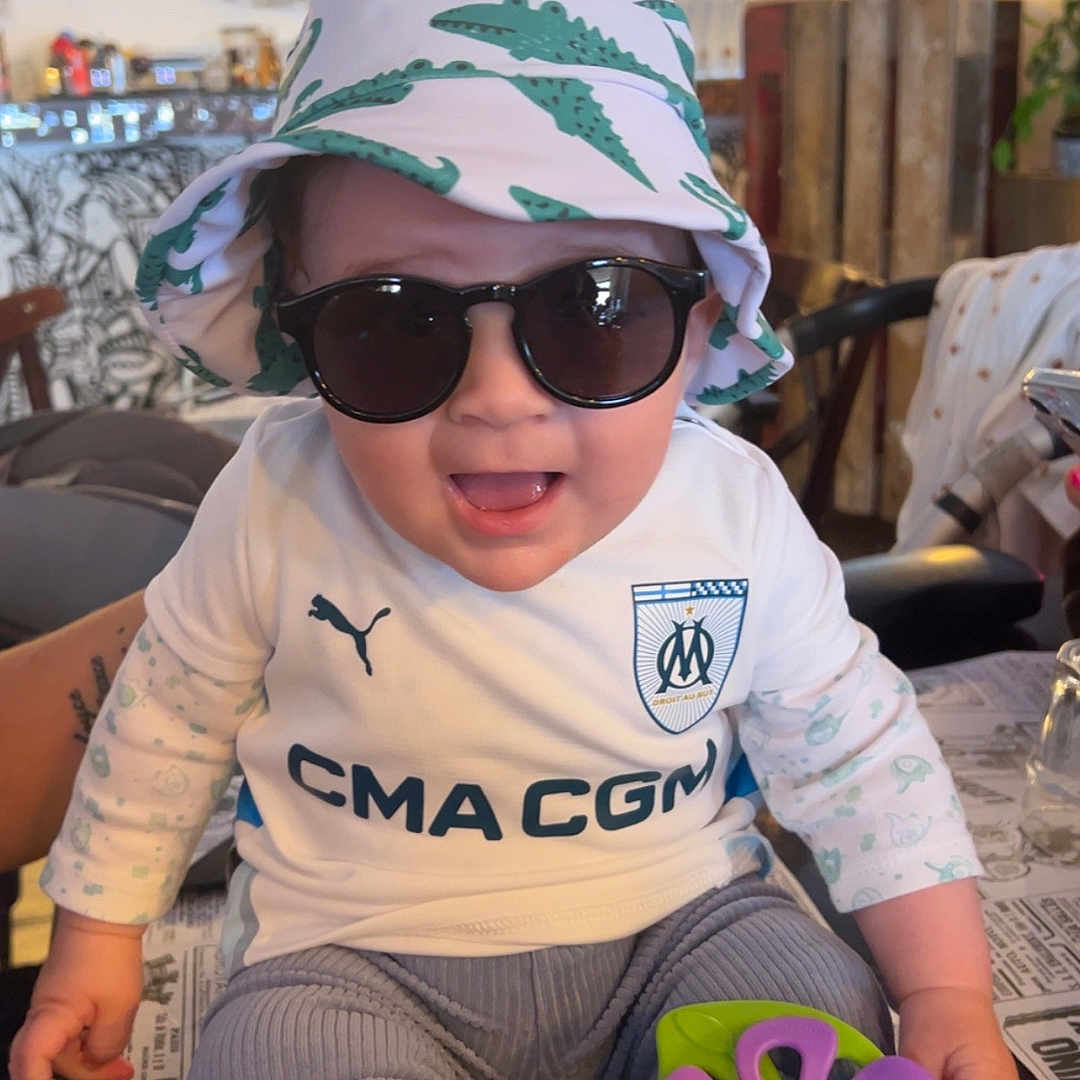 Nolan participe au concours pour gagner de l'argent avec cette photo : accessory, baby, bucket_hat, cafe, child, clothing, cute, fashion, happy, hat, indoor, long_sleeves, pants, person, seated, smiling, socks, sunglasses, table, toy