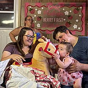Paisley is registered to the contest to win money with this photo: baby, child, woman, man, glasses, plush_toy, rocking_horse, birthday_party, gift_wrapping, gift, dress, indoor, smile, celebration, decorations, banner, family, happy, person, wood_paneling