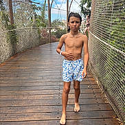 Djulian a rejoint le concours — aidez-le/la à gagner de superbes lots ! child, boy, bridge, wooden_deck, safety_net, fence, tropical_plants, trees, vacation, outdoor, barefoot, swim_shorts, patterned_shorts, necklace, wristband, walking, confused_expression, sunny_sky, person, background_child