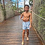 child, boy, bridge, wooden_deck, safety_net, fence, tropical_plants, trees, vacation, outdoor, barefoot, swim_shorts, patterned_shorts, necklace, wristband, walking, confused_expression, sunny_sky, person, background_child