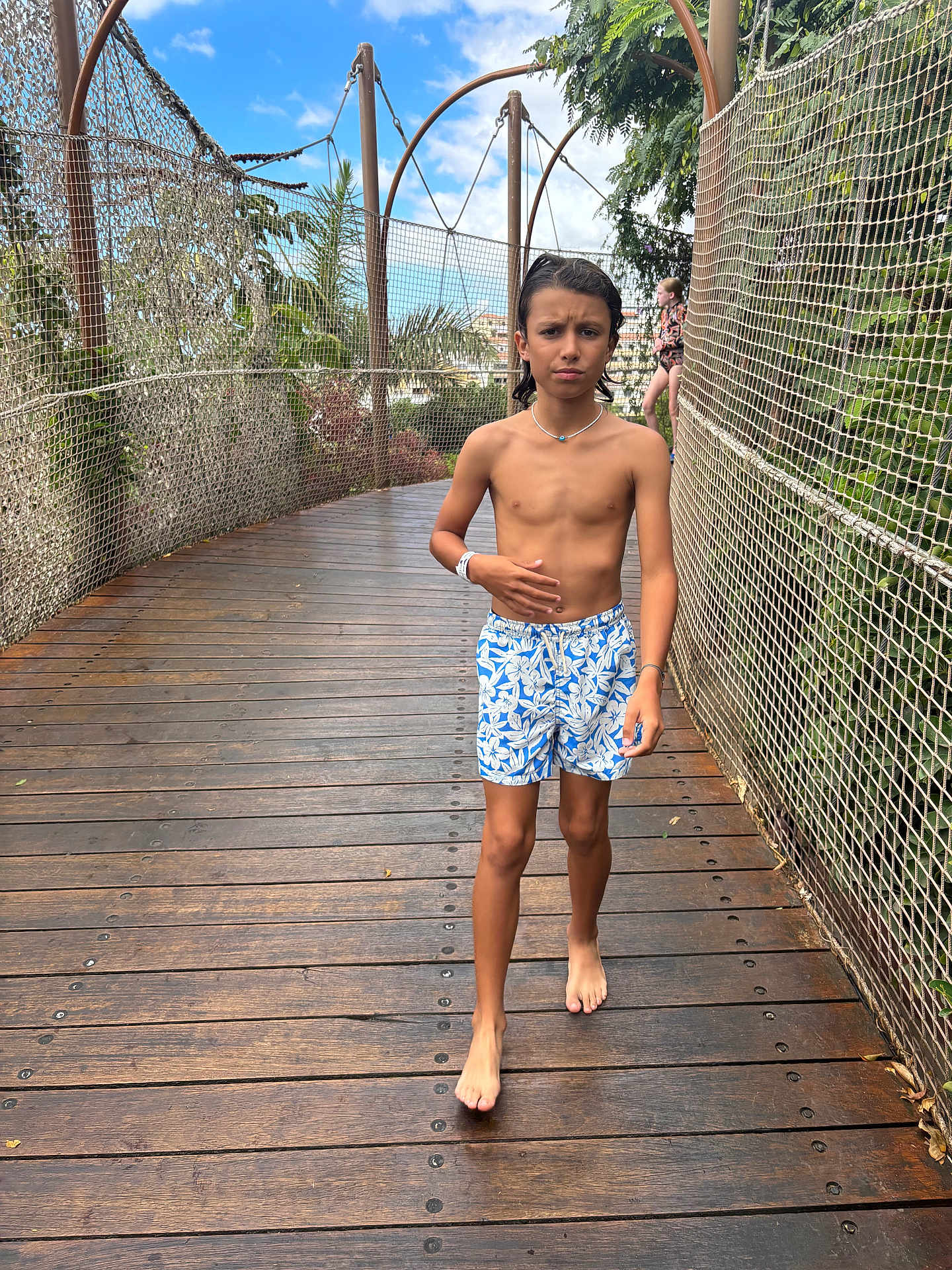 Djulian a rejoint le concours — aidez-le/la à gagner de superbes lots ! child, boy, bridge, wooden_deck, safety_net, fence, tropical_plants, trees, vacation, outdoor, barefoot, swim_shorts, patterned_shorts, necklace, wristband, walking, confused_expression, sunny_sky, person, background_child