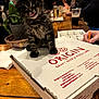Gandalf joined the competition — help win amazing prizes! kitten, cat, pizza_box, wooden_table, indoor, people, beverages, blurred_background, plant, hand, writing, menu, tattoo, casual, restaurant, night, curious, tabby, fur, tongue